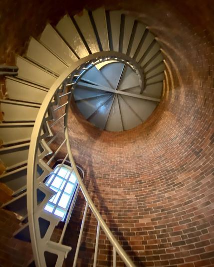 A dramatic upward view of a spiral staircase inside a brick lighthouse tower. The metal steps curve tightly around the cylindrical brick walls, leading up to a circular ceiling with a central support structure. A single window on the left lets in natural light, casting a soft glow on the warm red bricks and metal railings. The perspective emphasizes the staircase’s elegant, winding form as it disappears into the upper levels.