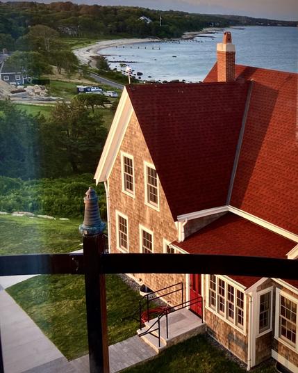 A scenic coastal view at sunset, captured from a high vantage point behind a black metal railing. Below, a charming shingled house with a steep red roof and bright red door is bathed in warm golden light. A paved walkway leads to the entrance, with a black metal handrail guiding the way. Beyond the house, a curving shoreline with a sandy beach and scattered rocks stretches along the calm blue ocean. A winding road follows the coastline, passing by trees, parked cars, and a few houses nestled in the lush green landscape.
