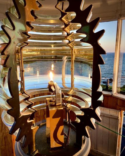 Close-up of a lighthouse Fresnel lens encasing a small, lit bulb. The lens, made of concentric glass rings and framed by black metal, magnifies the flame’s glow. Sunlight from a window behind reflects on the lens and the ocean beyond, casting warm golden hues across the scene. The sea and coastline are visible through the glass windows of the lighthouse.