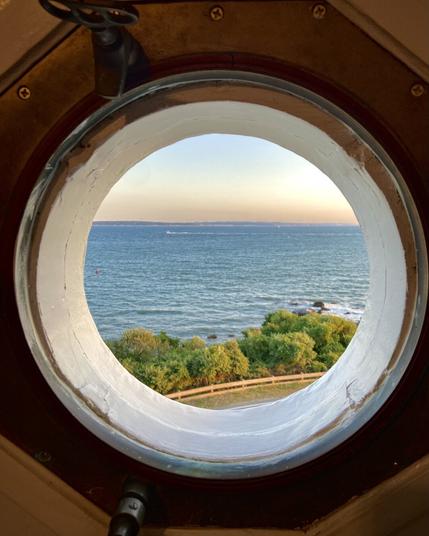 View through a round lighthouse porthole window set in a thick wall, looking out at a calm ocean under a clear sky during sunset. The horizon is tinged with soft pastel colors, and the foreground shows a strip of green trees and shrubs along a gently curving path near the rocky shoreline. The porthole frame and bolts are visible, adding a sense of enclosure and perspective.