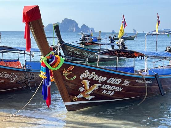 Bug eines am Sandstrand vertäuten Holzbootes. Das Boot ist am Bug mit Bändern und Girlanden in allen Farben geschmückt, dahinter ist es an der Bugwand mit einem Adler, lateinischer und thailändischer Schrift bemalt. Im Hintergrund sieht man das Meer mit weiteren Booten und einer hoch herausragenden, im Dunst nur noch unklar erkennbaren Insel.