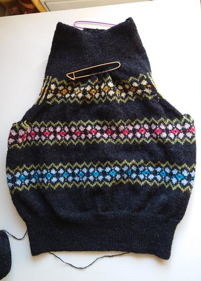 Shows a sleeveless black knitted top with three flower-like motif stripes. The bottom is blue, the middle is pink, and the top is yellow. The flowers are surrounded by a green zigzag border. It's a weird shape because the neck and sleeves still need to be cut out. It's lying on a white table.