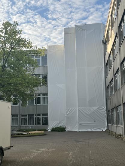 A building partially covered in white protective sheeting, surrounded by greenery and a paved area. The sky is partly cloudy, and there is a trailer in the foreground.