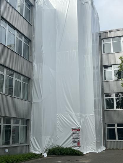 An exterior wall of a building is covered in white protective sheeting, indicating renovation or maintenance work. Nearby, there is a notice poster attached to the sheeting. The image shows a partially obscured view of adjacent windows and some greenery in the foreground