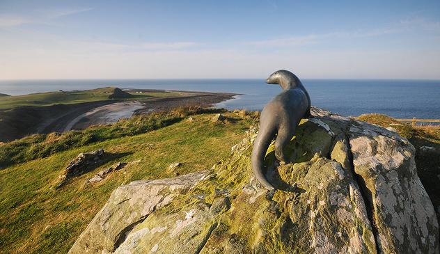 Photo of a bronze otter sculpture on a rocky outcrop, over looking a bay under a clear blue sky.