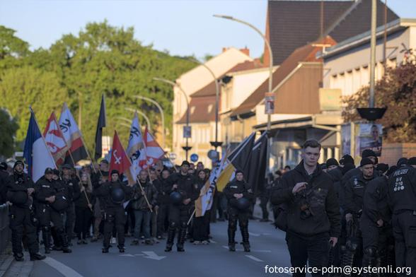 im vordergrund eine nazifotograf, der das "white power" symbol zeigt, im hintergrund der wartende neonaziaufmarsch