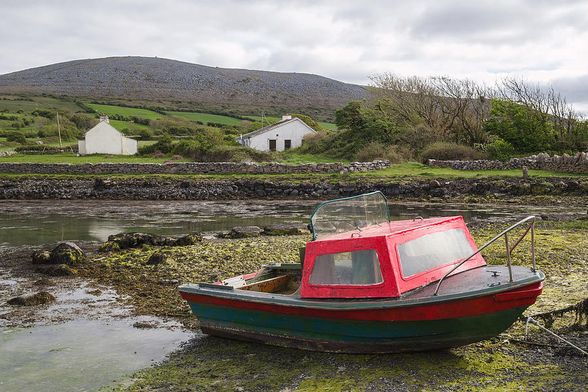 A small red and green angling boat is anchored at low tide on the Muckinish peninsula at Galway Bay in the Burren, County Clare, Ireland. The inlet is a well known spot for shore and sea angling. One of the Burren's stark gray karst limestone hills is seen in the background, with small green enclosed fields and two white houses filling the mid-ground.