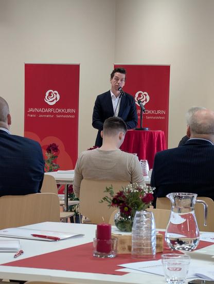 Kaare Dybvad Bek is talking into a microphone in front of a crowd sitting at tables inside. Behind him are two banners for the Faroese Social Democratic Party