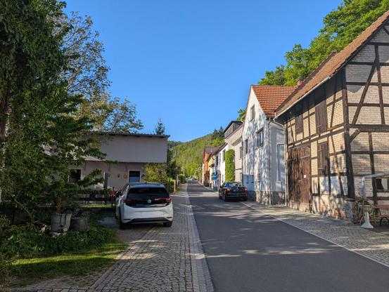 Photo of a small town street. On the right in the foreground is a half-timbered house. The street itself is asphalt and about 1 US lane wide; on each side there's a shoulder/sidewalk of stones. On the left an electric VW is parked. The sky's a beautiful cloudless blue. It's just rustic as heck here. If this photo had audio, you'd hear birds chirping.

Note: Number plates redacted using an "AI" infill method