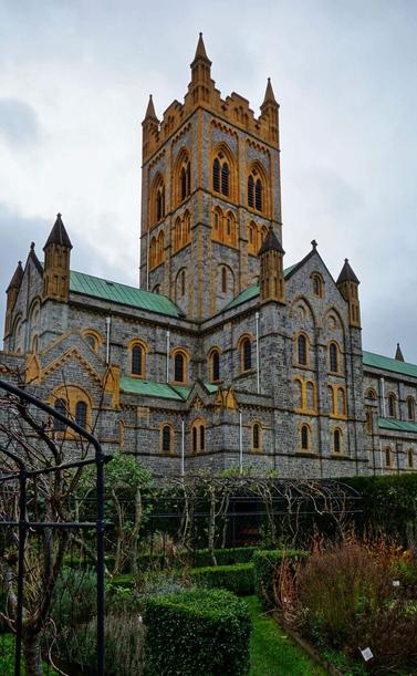 A low-angle shot shows a large, grey stone abbey with a prominent central tower under a cloudy sky. The tower and sections of the building feature yellow sandstone detailing around the windows and edges. A green roof covers parts of the abbey. In the foreground, a garden with low hedges, bare vines on a black metal frame, and various plants are visible, slightly out of focus.