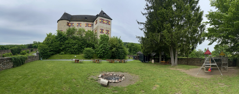 Burghof mit einer Wiese und einem Lagerfeuerplatz vorne und der Ritterburg umgeben von Bäumen hinten