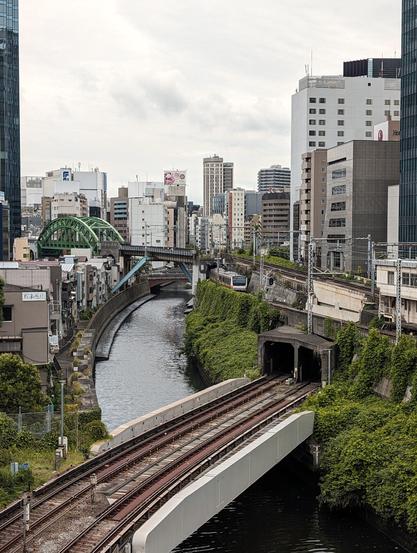 A train bridge over a bridge in the middle of Tokyo. The river is surrounded by high buildings.