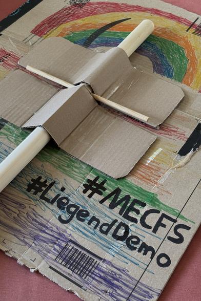 Ein Demo-Schild, Pappe am Holzstab, mit der Vorderseite nach unten liegend. Die Rückseite ist mit bunten Regenbögen bemalt und trägt die Hashtags #MECFS und #LiegendDemo in einer Ecke, etwa Handbreite Schriftgröße.