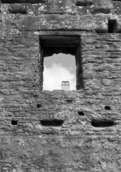 Black and white photograph looking up the stone wall of a ruined building. The viewpoint is from within the building, looking out an upper floor window. Through the window a portion of sky and the top of a chimney can be seen.
