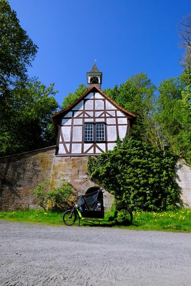 Photo of a neon-yellow cargobike in front of a building. The building has a massive sandstone base, a brown and white half-timbered facade, and a little turret on top. It is surrounded by trees.