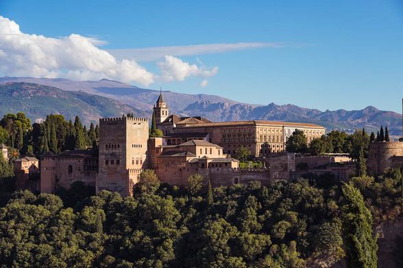 View of the Alhambra palace complex in Granada, Spain, with its reddish stone buildings set against forested hills and the Sierra Nevada mountains in the background, taken from a popular overlook at sunset.