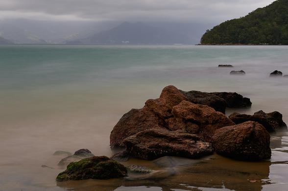 Upfront some reddish rocks with green algae, a milky turquoise sea and some mountains with green Atlantic rainforest on the right side in the back. The sky is completely covered with clouds.