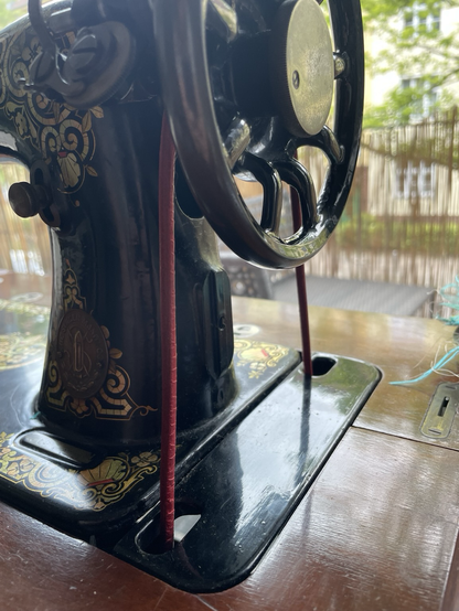 Close view from the right side of an old sewing machine. The balance wheel and a new red-ish leather belt are at the front.