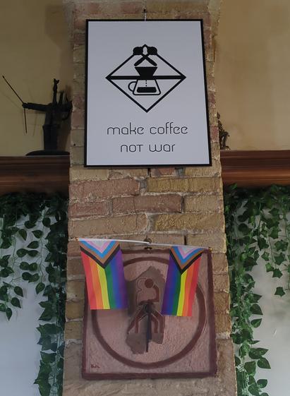 Photograph of a white sign showing the logo of the spot and a line stating "make coffee not war" underneath. Below there are two progress pride flags arranged in a v-shape.