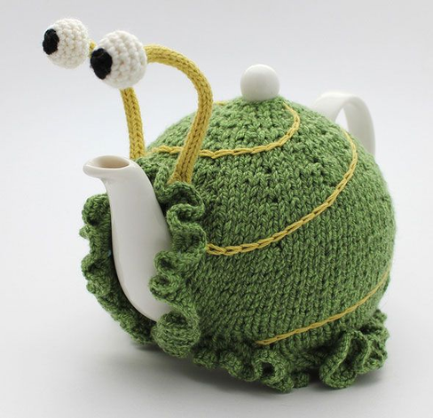 A white ceramic teapot wearing a green knitted tea cosy designed to look like a whimsical snail. The cosy features yellow lines mimicking a shell spiral, frilly edges around the base and spout, and two tall yellow stalks with crocheted eyes perched on top.