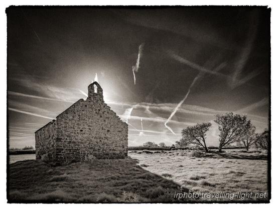 A toned black and white infrared photo of a ruined chapel, backlit by the sun against a blue sky with many aircraft contrails and wispy clouds.