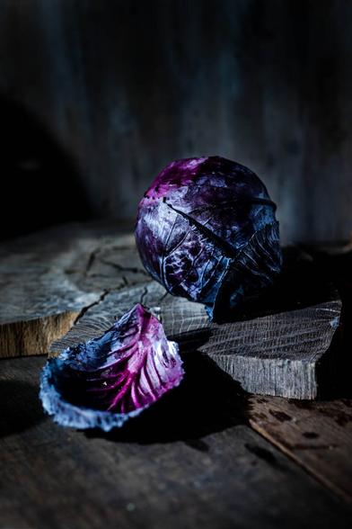 single red cabbage head on a wooden board in front of dark wooden background. A cabbage leaf in the front. The photo is dark and moody
