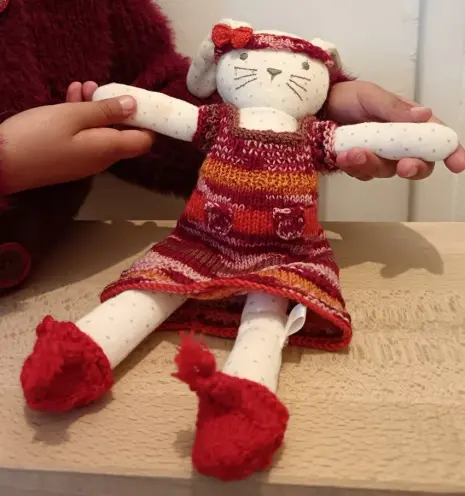 Lapin en tissu blanc à poids gris avec une robe en tricot dans les tons orange, rouge et rose. il a un bandeau de la même couleur avec un nœud orange et des chaussures rouges.