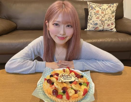 A woman with pastel pink hair is smiling at the camera while sitting behind a colorful fruit tart with a birthday message for “Hiromi.” She is seated in a cozy room with a brown leather sofa and a floral-patterned pillow in the background.