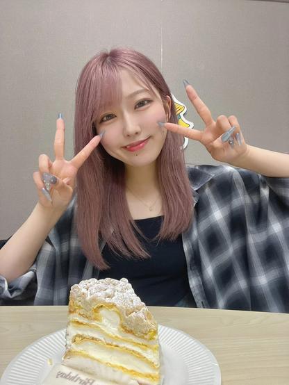 A cheerful woman with pink hair poses with a double peace sign, smiling brightly at the camera. In front of her is a slice of layered cream cake topped with powdered sugar on a white plate.