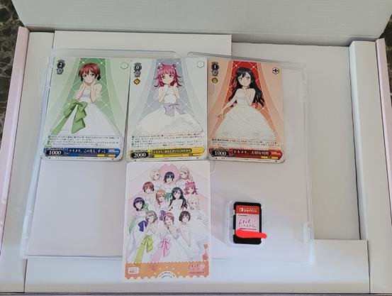 De game case van de Lovelive Nijigasaki visual novel voor Nintendo Switch opengespreid in de box editie

Zichtbaar zijn de game card, en 4 promo kaarten voor de trading card game

Waarvan 1 alle personages in een trouwjurk heeft, en de andere 3 elk apart een personage erop geprint hebben (Emma, Rina, Setsuna)