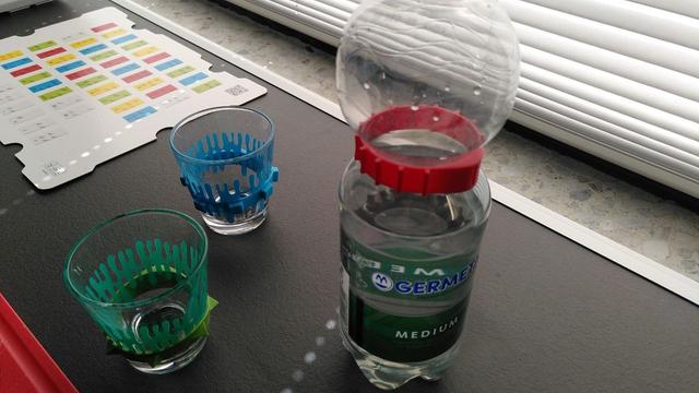 Eine PET-Wasserflasche und zwei Trinkgläser. 

Um die Gläser und die Wasserflasche sind Gummis gespannt, die reliefartige taktile Elemente beinhalten. Rillen, Spitzen oder Rechtecke. 

Der Gummiring um die Wasserflasche ist rot. Die Gummiringe um die Gläser sind grün und blau. 

Im Hintergrund liegt eine Referenztafel zu den Braille-Steinen von LEGO.