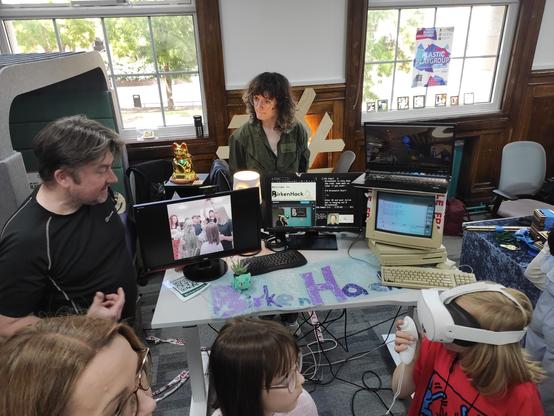 An overhead view of the Birkenhack stall at Wirral Makefest kids and adults are gathered around, one kid is wearing a VR headset and interacting over the internet with Julian, who is wearing a headset in Birkenhack