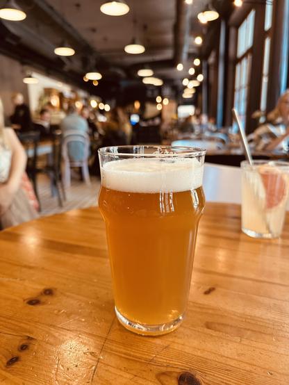 A pint glass of Anarkist IPA sits on a wooden table in Andalūzijas Suns, a lively bar in Rīga. The beer has a hazy golden-orange color with a frothy white head. In the background, the bar’s spacious, industrial-style interior is softly lit with hanging lamps, and patrons are enjoying drinks and conversation, creating a warm and bustling atmosphere.