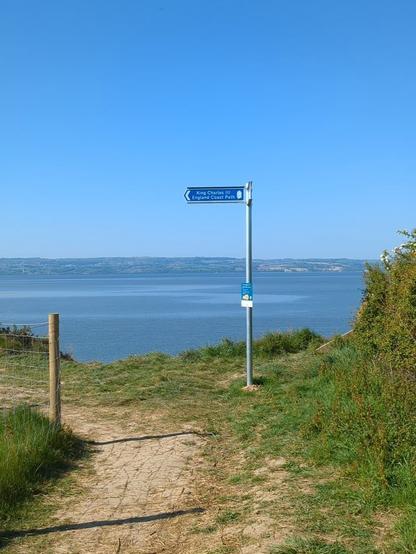 A dirt path leads to a scenic estuary view, with a signpost under a clear blue sky.