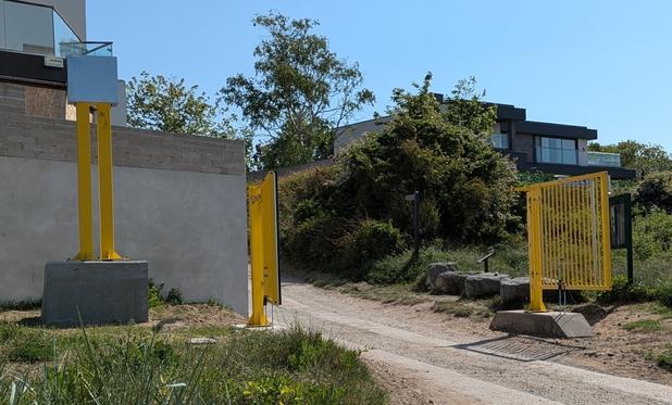 Yellow gate positioned along a pathway, with buildings and greenery in the background.