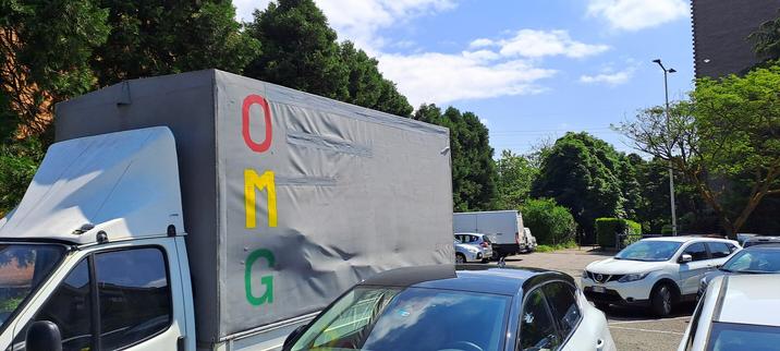 The image shows a parking lot with several vehicles. In the foreground, there is a white truck with a gray cargo area. The letters "OMG" are prominently displayed vertically on the side of the cargo area, with "O" in red, "M" in yellow, and "G" in green. The truck is parked among other vehicles, including cars and another truck in the background. The parking lot is surrounded by green trees and a clear blue sky with some clouds. The scene appears to be in a sunny, outdoor setting, possibly in a suburban or urban area.