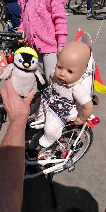 Auf einem Kinderrad ist ein Sitz montiert in dem eine Puppe angeschnallt sitzt. Archy sitzt für ein Foto daneben.