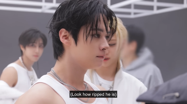 Lee Know in a white sleeveless top. Felix stands behind him and looks at his arms and back. Caption: (Look how ripped he is)