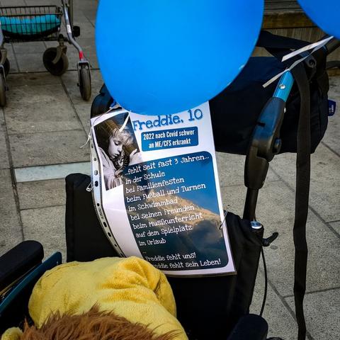 Das Foto zeigt einen leeren Kinderrollstuhl. Stattdessen sitzt ein Plüsch-Löwe darin. Zwei blaue Luftballons sind hinten am Rollstuhl befestigt. An der Lehne des Rollstuhls, ist ein DIN A4-Blatt mit Informationen angeheftet, das ein Foto eines liegenden/schlafenden Kindes (Junge 10 Jahre) zeigt. folgender Text:

Freddie. 10. 2022 nach Covid schwer an ME/CFS erkrankt. 

...fehlt seit fast 3 Jahren... 
in der Schule
bei Familienfesten
beim Fußball und Turnen
im Schwimmbad
bei seinen Freunden
beim Musikunterricht
auf dem Spielplatz
im Urlaub
...
Freddie fehlt uns!
Freddie fehlt sein Leben!