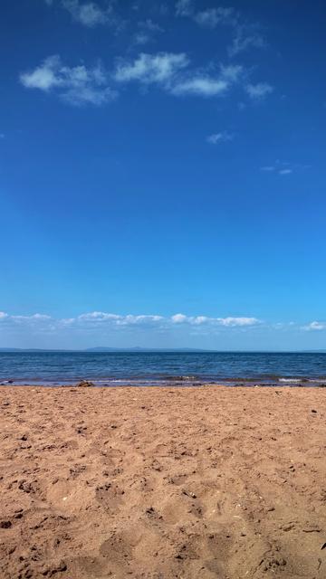 A clear view from a sandy beach looking out toward a calm blue sea under a mostly clear sky with scattered clouds.