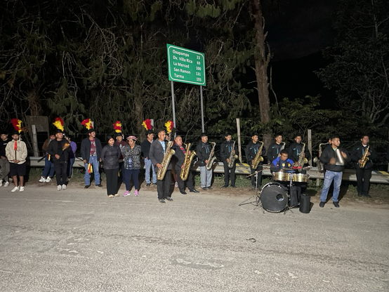 Bailantes de chunginada en la noche con musicantes de la orquesta folclorica. Adelante Saxo bajo por la derecha otros saxos y batería. En el fondo oscuro se ven arboles y un letrero de carretera.