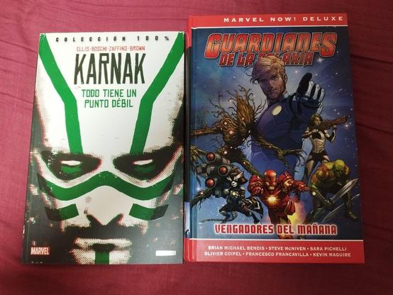 Dos cómics colocados uno al lado del otro sobre una superficie roja. El cómic de la izquierda se titula 'Karnak: Todo Tiene Un Punto Débil' de la colección '100%'. En la portada se muestra un primer plano de un rostro con marcas verdes y blancas, y en la parte superior aparecen los nombres Ellis, Boschi, Zaffino y Brown. El cómic de la derecha se titula 'Guardianes de la Galaxia: Vengadores del Mañana' de la serie 'Marvel Now! Deluxe'. Su portada presenta a varios personajes, incluidos Groot, Rocket Raccoon, Iron Man y otros, con un fondo espacial. Los nombres Brian Michael Bendis, Steve McNiven, Sara Pichelli, Olivier Coipel, Francesco Francavilla y Kevin Maguire aparecen en la parte inferior.