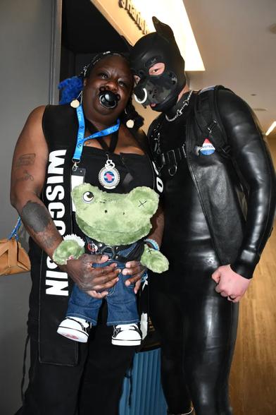 Two IPTC attendees stand side by side in an exhibition hall. On the right, a ponyhound is clad in a Furrjoi black silicone body suit: a neoprene Mr. S puppy hood with a white nose ring, a chain collar with a snapple bit, a black harness, and a leather bar vest, accented by an “I Voted” sticker on their chest. 

On the left, a smiling Black lady wears a sleeveless black top, a leather sash emblazoned with “Wisconsin Bootblack,” chunky silver lip jewelry, cobalt blue braids, and holds a large plush frog dressed in denim pants and sneakers. Corridor lighting highlights the textures of neoprene, leather, metal, and plush fabric. 

Source: https://windycitytimes.com/2025/04/09/pups-and-trainers-give-their-all-at-chicago-competition/
