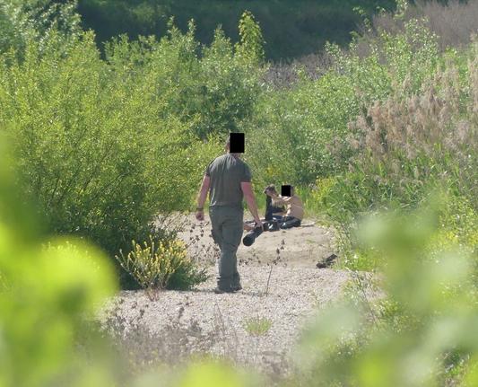 Mann mit Kamera und großem Objektiv läuft auf Kiesbank, weiter hinten ein Paar, halbnackt, kleiden sich gerade sitzend an, Fotograf läuft auf sie zu, wir sehen ihn von hinten
