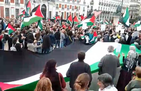 Enorme bandera palestina llevada en horizontal por personas alrededor de ella. Muchedumbre con muchas banderas palestinas, en Madrid.