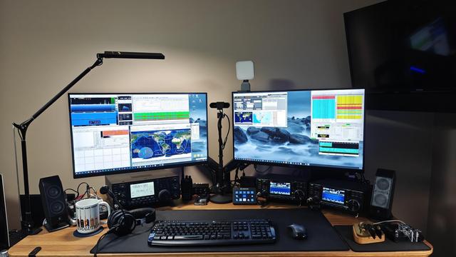 Desk with a IC-9100 to the left and a IC-9700 and IC-7300 to the right. Above them are two 27" panels. In front of the radios are a keyboard, mouse, and a headset.