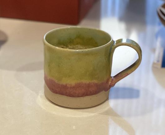 A medium coffee mug sits, handle-right, on a white counter.  The mug is glazed a fairly bright yellowish green (mottled) in the upper two thirds, followed by a drippish band of magenta, ending with the pale (whitish) clay at the bottom.  On the whole, it feels like a bit of sunshine in the morning!