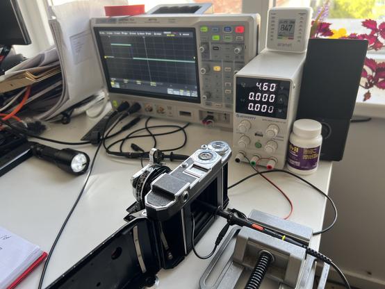 Overview of the setup with camera in the middle, a flashligth shines into the lens, a photodiode with resistor is held into the back. It is powered by a small laboratoy power supply. A digital storage oscilloscope showes the time of the shutter.