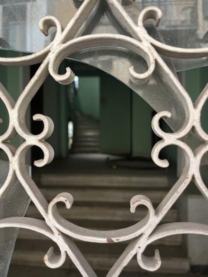 It’s a close up on a door of glass and wrought iron, the pattern a diamond embellished with curls. The metal has been painted white and the glass is broken. Through the broken glass plain white marble steps lead to  the lobby of an apartment building, painted turquoise. There’s an elevator ansd also stairs leading up.