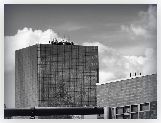 Black and white photo.
cloudy sky behind skyscraper, smaller brick building on the right. The clouds are reflecting in the windows of the skyscraper.